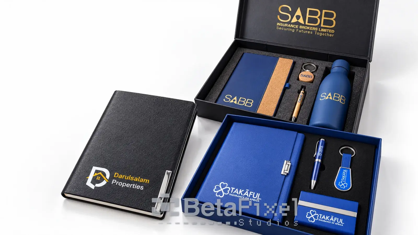 Two executive corporate gift boxes with branded notebooks, pens, keyrings, a water bottle and a card holder, beside a leather notebook with a logo