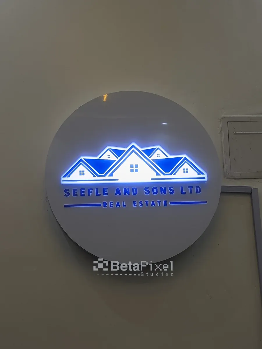 Round grey wall sign with a blue LED-lit rooftop logo for a real estate company, installed on an office wall