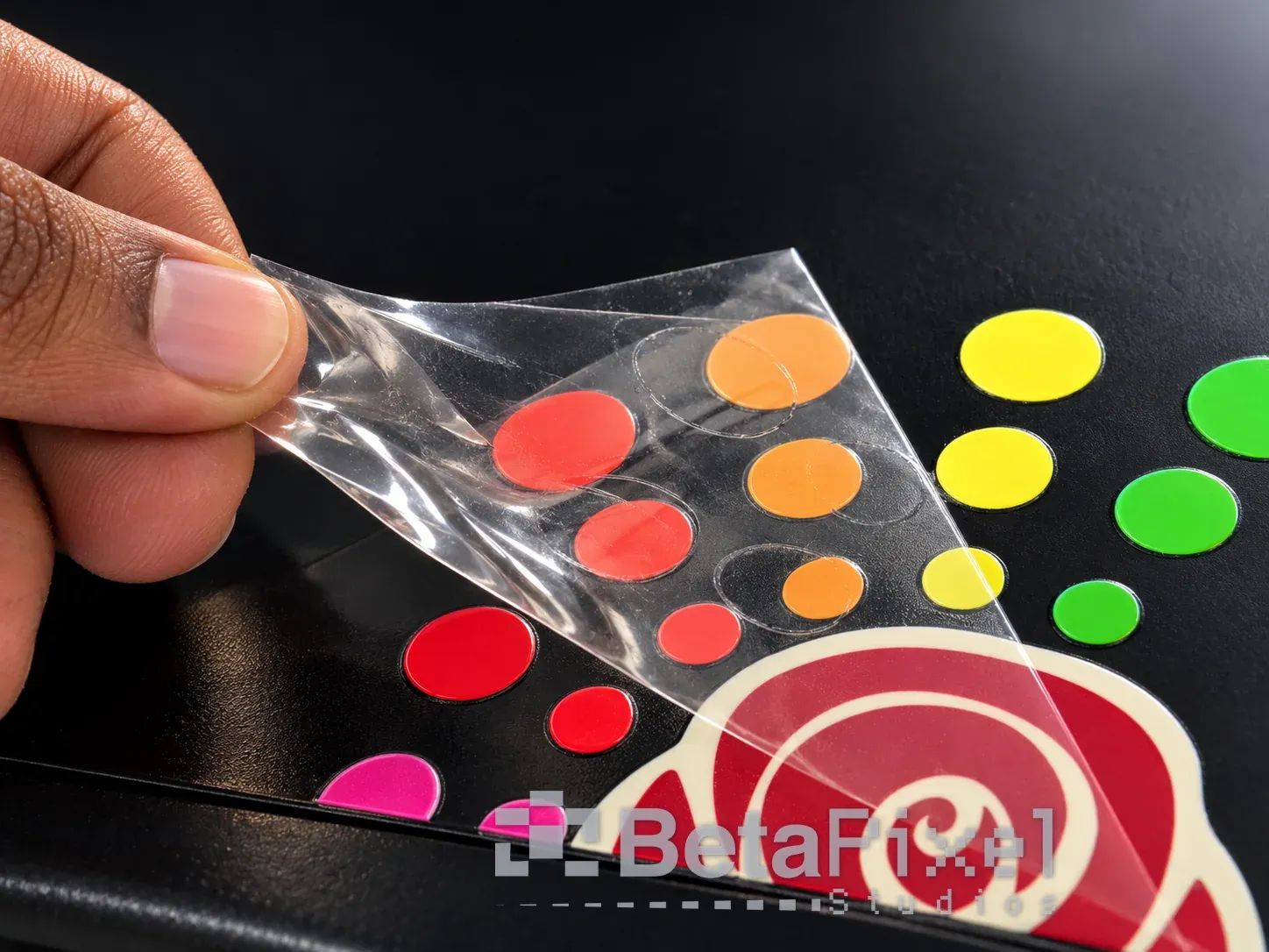 Hand peeling the clear film away from colourful UV DTF circle and rose transfers on a black surface, leaving only the printed design